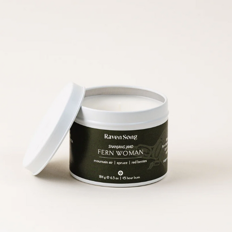 A white candle container with a black label that reads 'RavenSong' and 'FERN WOMAN'. The lid is off to the side, revealing a white candle inside.