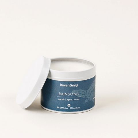 A Ravensong branded candle with the name Rainsong, featuring a white candle in a container with a label that includes amber, jasmine, and tonka scents.