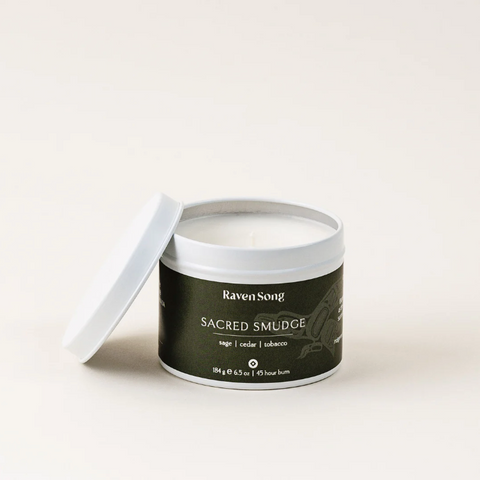 A white candle in a metal container with a label reading 'Sacred Smudge' on a light background.