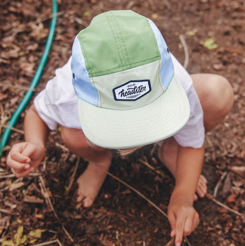 Child wearing a green and white cap with a brand logo, sitting on the ground.