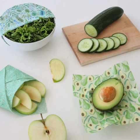 Green reusable food wraps with sliced cucumbers, avocado, and apples on a white background