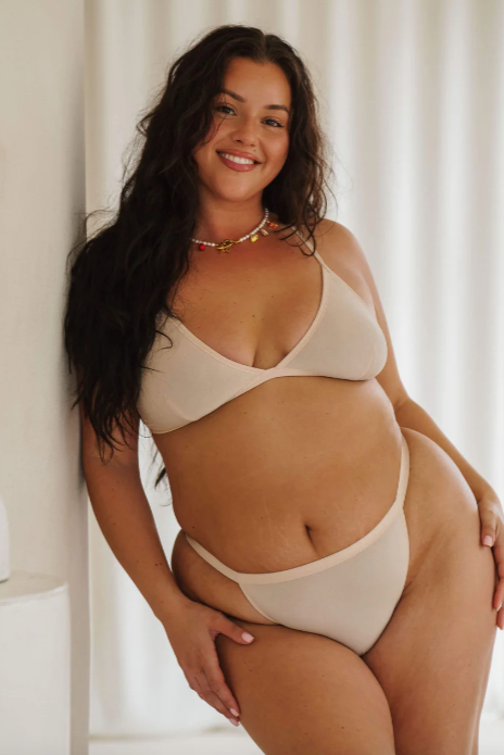 Woman wearing a beige bikini sitting against a white curtain.