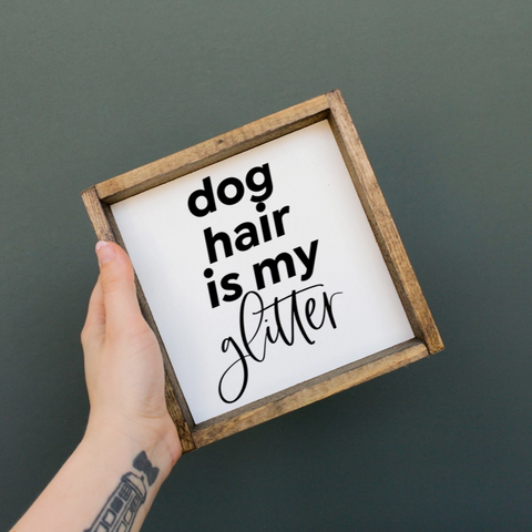 Hand holding a framed sign with 'dog hair is my glitter' text on a dark background