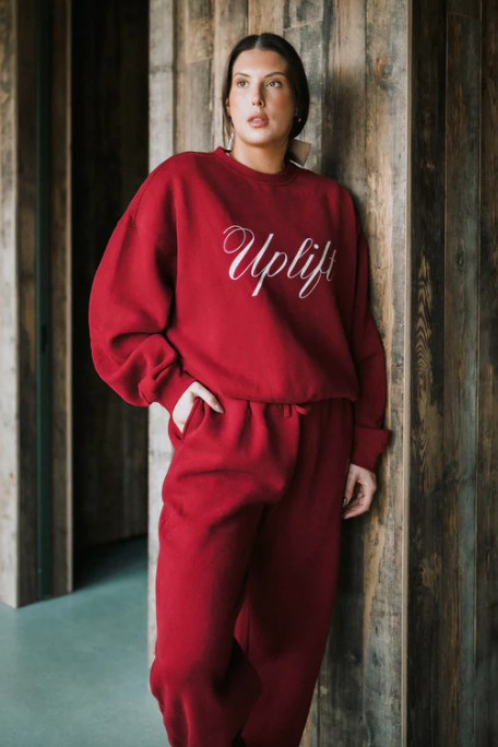 Person wearing a red 'Uplift' sweatshirt and pants against a wooden wall.