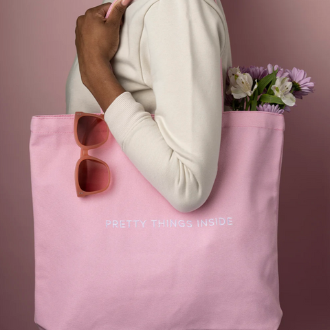 Person holding a pink tote bag with 'Pretty Things Inside' text, sunglasses, and flowers against a pink background.