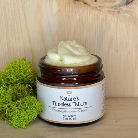 A jar of 'Nature's Timeless Tallow' moisturizer on a wooden surface, with a label that includes the product name and the claim of being 'Toxin Free'. The jar is accompanied by a small amount of product visible on a green moss pad.