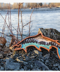 A fish-shaped wooden wall art piece featuring a colorful landscape design, displayed on a tree stump in an outdoor river setting.