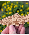 A hand holds a laser-engraved wooden fish-shaped plaque featuring a mountain, trees, a fishing boat, and the name 'Campbell Riverr' against a blurred background of yellow flowers.