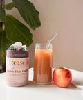 Tea container labeled 'teast' next to a glass of juice and an apple on a surface with a plant in the background.