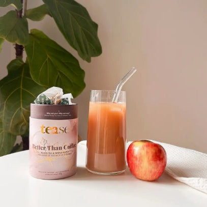 Tea container labeled 'teast' next to a glass of juice and an apple on a surface with a plant in the background.