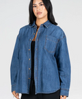 A woman modeling an oversized blue denim button-up shirt with a chest pocket, worn open over a black top and black pants, against a light gray studio background.
