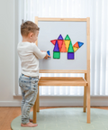 A young child stands barefoot in front of a wooden magnetic activity board, placing colorful geometric shapes onto the white writing surface in a bright room with sheer curtains and a plant.