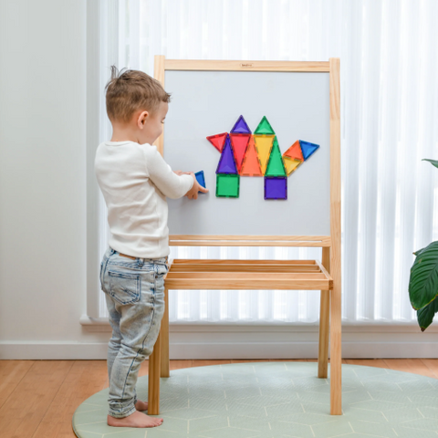 A young child stands barefoot in front of a wooden magnetic activity board, placing colorful geometric shapes onto the white writing surface in a bright room with sheer curtains and a plant.
