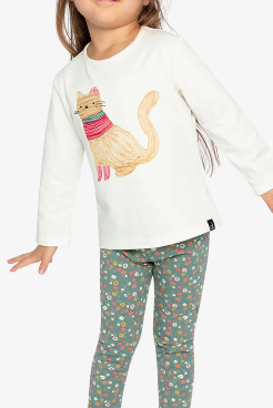 A young girl modeling a white long-sleeve top with a colorful cat graphic and green patterned leggings, standing against a light gray studio background. A small label in the top right reads 'Coton biologique'.
