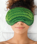 A person lying down wearing a green watermelon-patterned eye mask over their eyes, with white towels behind them.