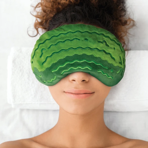 A person lying down wearing a green watermelon-patterned eye mask over their eyes, with white towels behind them.