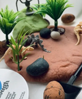 Play dough landscape with plants, rocks, and a tool on a white surface