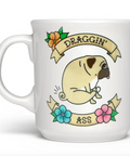 A white ceramic coffee mug featuring a cartoon pug sitting on a banner that reads 'DRAGGIN ASS' with decorative flowers, shown on a white background with a Fred store header and macOS dock visible.