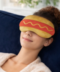A woman reclining on a dark blue couch wearing a plush hot dog sleep mask.