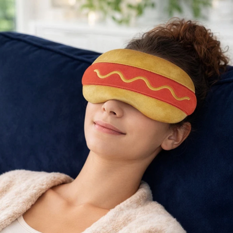 A woman reclining on a dark blue couch wearing a plush hot dog sleep mask.