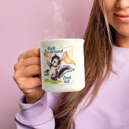 A person in a lavender sweatshirt holds a white ceramic mug with a cartoon skunk illustration and the text 'fuck around find out' against a solid pink background.