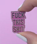 Pink and black enamel pin with text held by a hand against a pink background