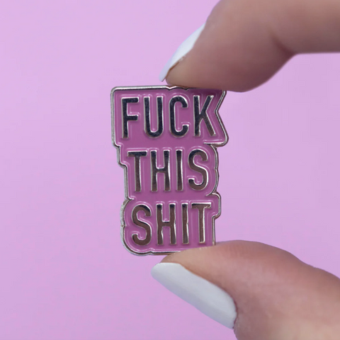Pink and black enamel pin with text held by a hand against a pink background