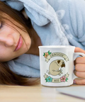 A woman in a light blue hoodie rests her head on a table while holding a white ceramic mug printed with a cartoon pug and the words 'DRAGGIN' ASS'. The scene appears to be a lifestyle product photo, but the right side shows an e-commerce webpage interface with product text and a macOS dock at the bottom, making this a mixed/other listing-style composite rather than a clean product shot.