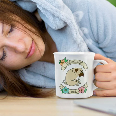 A woman in a light blue hoodie rests her head on a table while holding a white ceramic mug printed with a cartoon pug and the words 'DRAGGIN' ASS'. The scene appears to be a lifestyle product photo, but the right side shows an e-commerce webpage interface with product text and a macOS dock at the bottom, making this a mixed/other listing-style composite rather than a clean product shot.