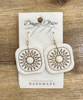 White earrings with gold designs on a 'Dayna Olson' card, placed on a wooden surface.