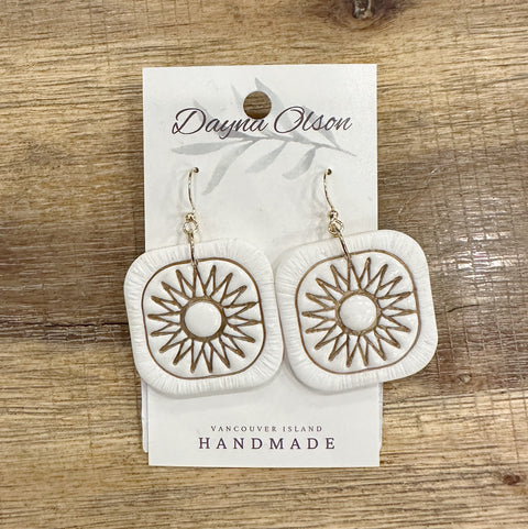 White earrings with gold designs on a 'Dayna Olson' card, placed on a wooden surface.