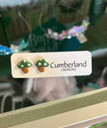 Mushroom-shaped earrings on a display card with 'Cumberland Creations' branding, reflected in a glass surface.