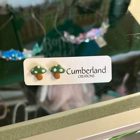 Mushroom-shaped earrings on a display card with 'Cumberland Creations' branding, reflected in a glass surface.
