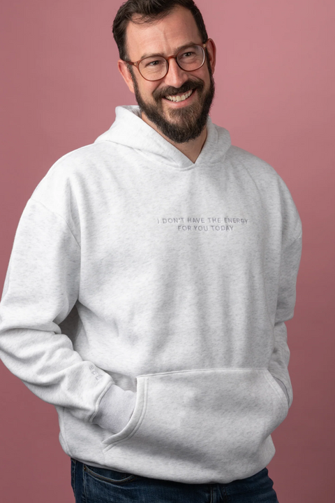 A smiling man with glasses and a beard wearing a light gray heather hoodie with the text 'I DON'T HAVE THE ENERGY FOR YOU TO TRY' printed on the chest, posed against a solid dusty pink background.