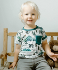 Child sitting on a wooden chair with a teddy bear, wearing a shirt with nature-themed design.