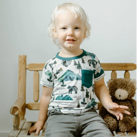 Child sitting on a wooden chair with a teddy bear, wearing a shirt with nature-themed design.