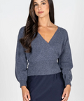 A woman modeling a slate blue-gray knit sweater with a V-neck and long sleeves, paired with a dark navy skirt, against a plain light gray studio background.