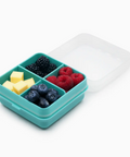 A teal silicone compartment lunch box with a translucent white lid, shown open and filled with blueberries, blackberries, raspberries, and pineapple chunks on a white background.