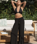 Woman in black bikini and wide-leg pants standing outdoors with greenery and furniture in the background