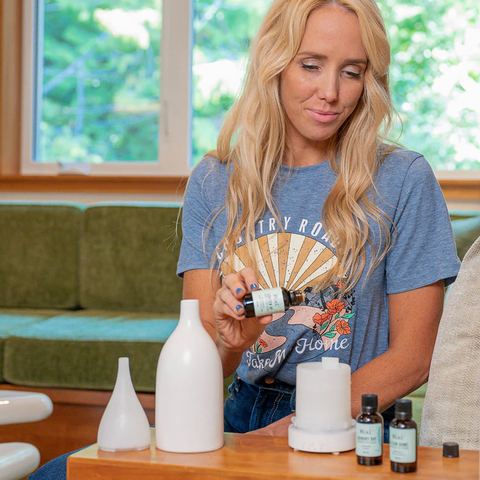 Woman holding a bottle of essential oils in a home setting with white bottles and a wooden table.