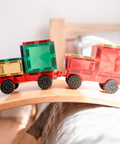 Two toy cargo trucks built from translucent magnetic building blocks and black wheels, resting on a curved wooden board in a softly blurred indoor setting.