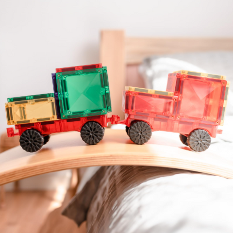 Two toy cargo trucks built from translucent magnetic building blocks and black wheels, resting on a curved wooden board in a softly blurred indoor setting.
