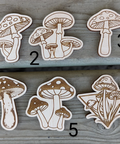 Six laser-cut wooden mushroom stickers or decals numbered 1 through 6, arranged on a weathered gray wooden surface. The mushrooms are carved from light wood with dark brown engraved details, featuring various mushroom designs including single, double, and triple clusters.
