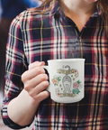 A person wearing a navy, red, and yellow plaid flannel shirt holds a white ceramic mug with a cute cartoon cat illustration and the text 'MY CAT IS A CAT' wrapped around it. The person is also holding a tablet in a softly blurred indoor setting.