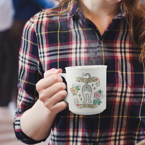 A person wearing a navy, red, and yellow plaid flannel shirt holds a white ceramic mug with a cute cartoon cat illustration and the text 'MY CAT IS A CAT' wrapped around it. The person is also holding a tablet in a softly blurred indoor setting.