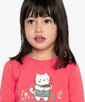 A young girl with dark hair wearing a bright pink long-sleeve shirt with a cartoon cat graphic and pink floral pants, photographed against a light gray background.