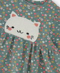 A close-up of a children's long-sleeve dress in sage green cotton fabric with a small-scale multicolor floral print. The dress features a gathered neckline and a white felt patch of a cat's face on the chest.