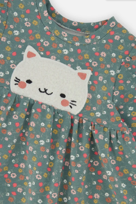 A close-up of a children's long-sleeve dress in sage green cotton fabric with a small-scale multicolor floral print. The dress features a gathered neckline and a white felt patch of a cat's face on the chest.