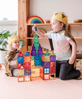 Two children wearing gold paper crowns and gold capes sit on a pink patterned rug in a bright playroom, building a colorful magnetic tile castle together.
