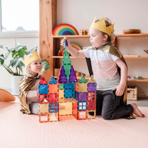 Two children wearing gold paper crowns and gold capes sit on a pink patterned rug in a bright playroom, building a colorful magnetic tile castle together.